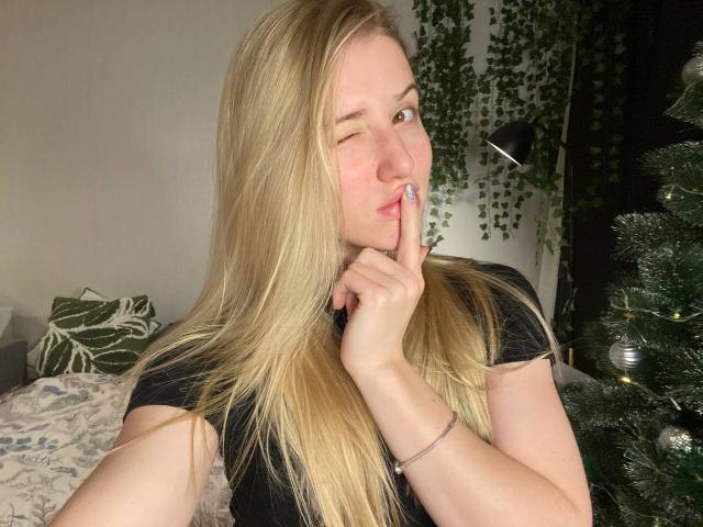 ArianaMur, blonde, winking, with finger to lips, black t-shirt, long hair, small nose ring, silver bracelet, Christmas tree to right, green plant hanging in background.