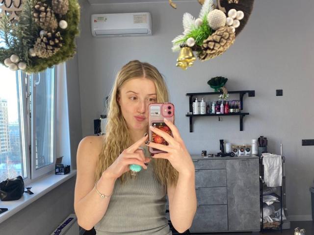 ArianaMur with blonde hair, wearing a gray tank top, holding a pink phone with a red case, taking a mirror selfie, showing her small tits, in a bright room.