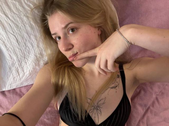 ArianaMur with blonde hair, pouty lips, and black pinstriped bra, holding her finger to her mouth, showing a flower tattoo on her chest. Pink bed and white pillow.