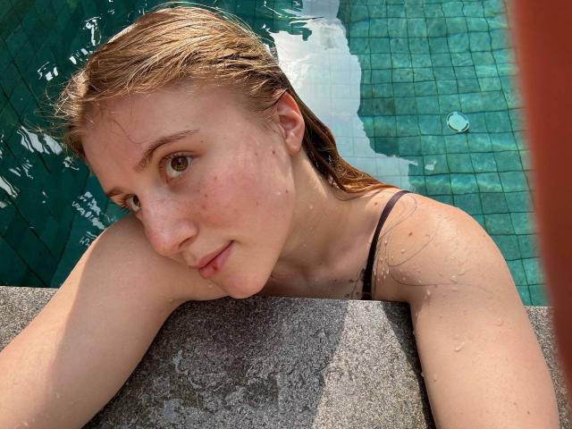 ArianaMur with wet blonde hair, green eyes, and freckles, leaning on a pool edge, wearing a black spaghetti strap, with water droplets on her skin.
