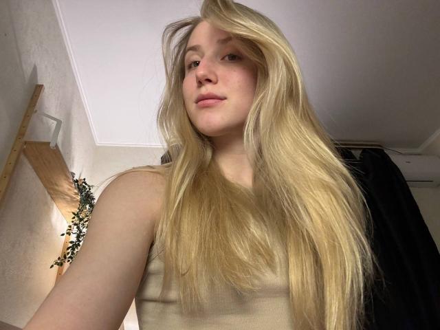 ArianaMur with long blonde hair, pale skin, and pink lips, wearing a beige tank top, showing her tits slightly, in a low-angle selfie.
