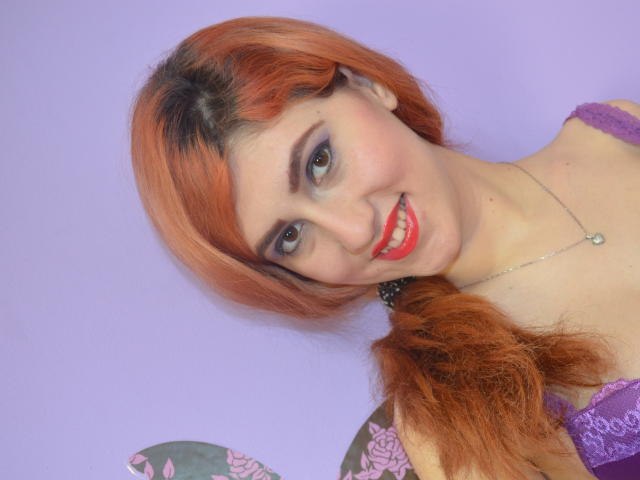 WonderPearl avec des cheveux roux, un eye-liner violet, du rouge à lèvres rouge, souriante, portant de la dentelle violette, un collier et une queue de cheval, sur un fond violet. Seins visibles sous le haut en dentelle..