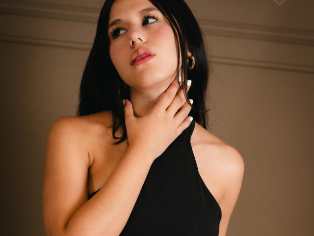 AmbarStoesselmml with dark hair, wearing a black halter top, touching her neck, looking up with blue eyes, pink lips, and white nails, in a soft-lit, brown-walled room.