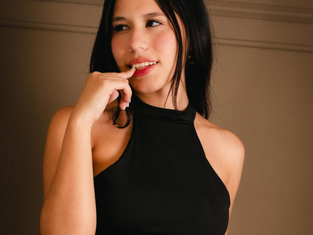 AmbarStoesselmml with black hair, fair skin, and red lipstick, wearing a black halter top, biting her finger, smiling, showing small tits and a cute nose.
