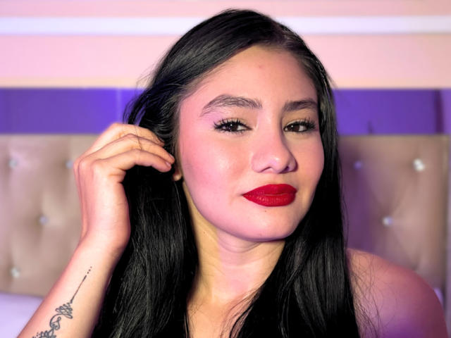 LailaTurbay has black hair, red lipstick, and a tattoo on her right wrist. She's touching her hair with a slight smile. Her tits are not visible.