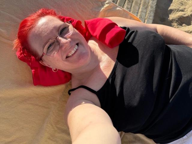 MrsEssen7471-hot with red hair, glasses, black tank top, and red pillow, smiling, lying on yellow blanket, sunlight on her face, showing her small tits and white pants.