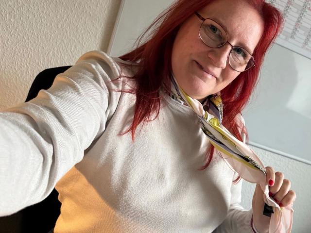 MrsEssen7471-hot with red hair, glasses, and white sweater, holding a yellow and black tie, smiling at the camera, with a white wall and calendar in the background.