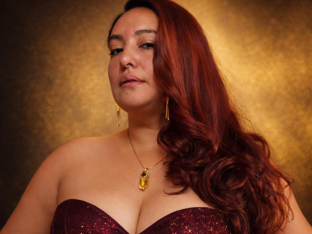 A TerraMegaBoobs tem cabelo ruivo, brincos de ouro e um colar de ouro com um pingente. Ela está usando uma blusa brilhante sem alças, que mostra seus seios grandes e brilhantes. Ela parece confiante..