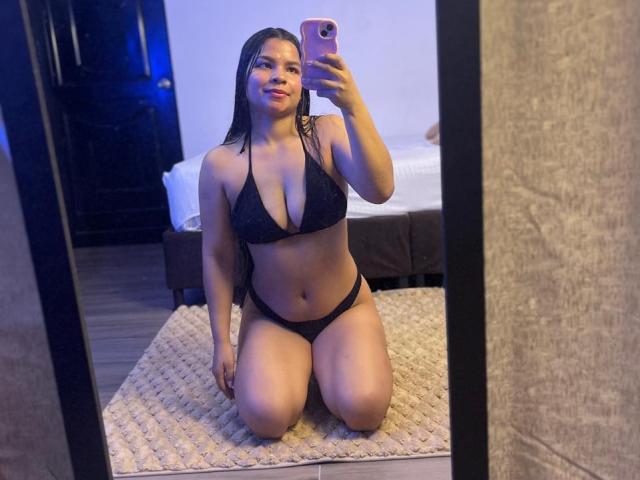 AmaiaSaenz kneeling in front of a mirror, wearing a black bikini, showing her tits and pussy, holding a pink phone, with a slight smile on her face.