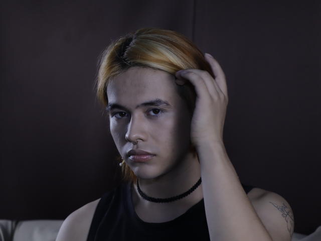 BenjiVampire with light brown hair, nose ring, black tank top, black choker, tattoo on right arm, hand in hair, serious expression, soft lighting, dark background, slightly bluish tone.