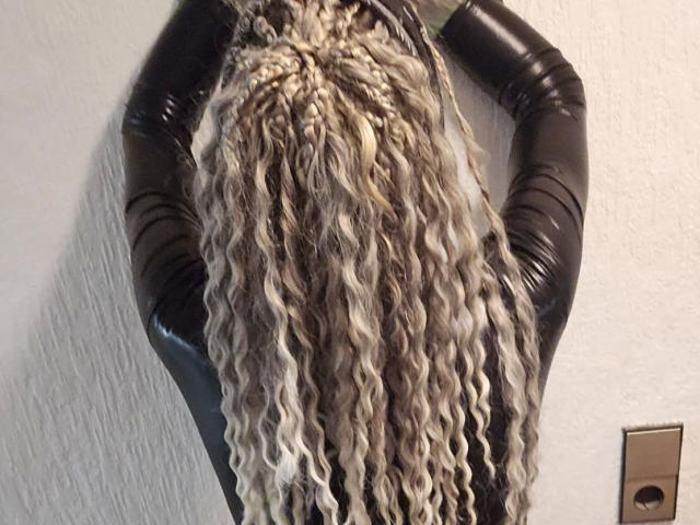 Chrissy-cupcakes-hot's blonde, curly, wavy hair covers their black leather arms, standing against a white wall. A brass electrical outlet is visible in the lower right.