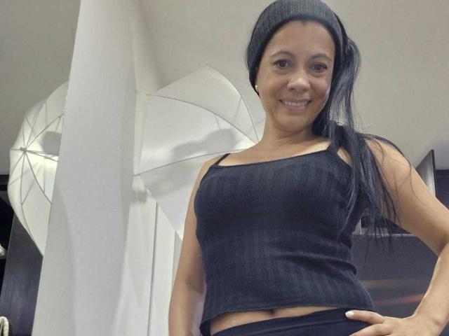 MelissaBaud with long black hair, wearing a black beanie, black tank top, and black pants, smiling, showing her small tits and a hint of her belly button.