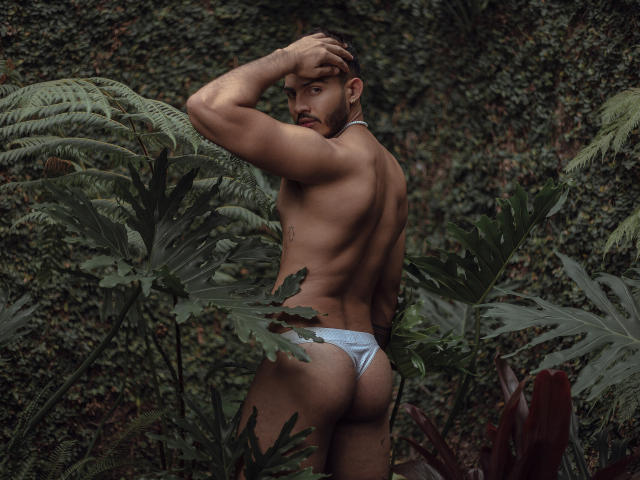 KlinsmannCampbell, met een gespierd, bruin lichaam, draagt een strak, wit stringbroekje, waarbij een rond, stevig achterwerk zichtbaar is. Hij staat in groene begroeiing, kijkt om en heeft een hand op zijn hoofd..