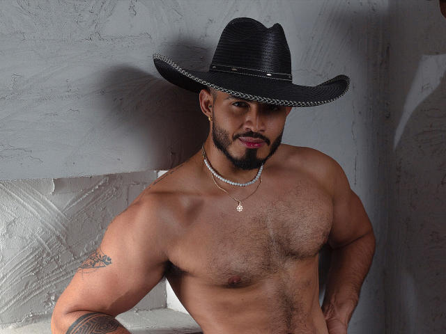 Muskulöser KlinsmannCampbell mit einem schwarzen Cowboyhut, Brusthaar, einem Tattoo am linken Arm, einer Halskette und Bart, steht oben ohne da, blickt mit einem leichten Lächeln in die Kamera..
