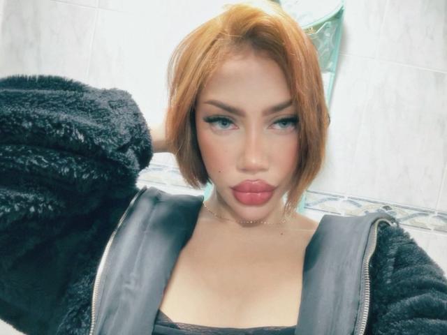 MelanieCollins has short red hair, black fuzzy jacket, and puffy red lips. She's looking at the camera with a slight pout, wearing a black top and choker.