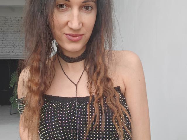 BanyReid with long, wavy brown hair, wearing a black polka-dot strap top, black choker, and necklace, showing her boobs, with a subtle smile and pink lipstick.