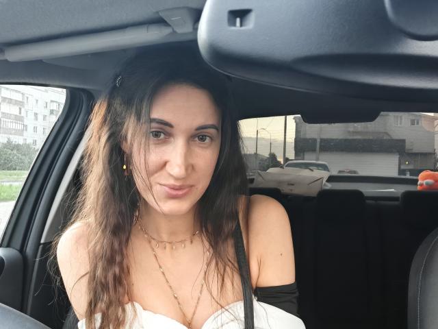 BanyReid with wavy brown hair, small gold earrings, and a white off-shoulder top, sitting in a car, smiling softly, showing small tits, and a delicate necklace.