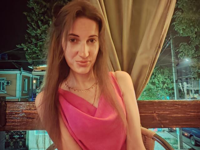 BanyReid with long brown hair, pink dress, and gold necklace, pouting slightly, sitting at a wooden table with green curtains, looking at the camera, night outside.