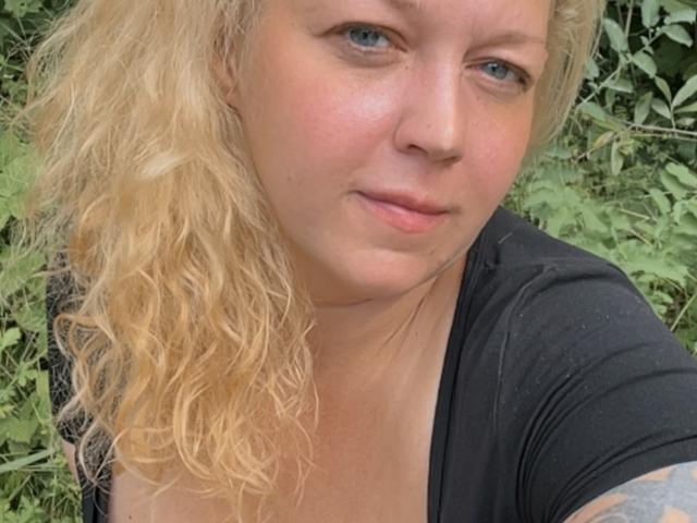 JenniferCarmen with wavy blonde hair, blue eyes, and black top, showing a subtle smile, her tits slightly visible, standing in greenery. Her tattooed arm is partially seen.