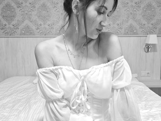 BanyReid with off-shoulder white dress, puffy sleeves, and ribbons, wearing long earrings and necklace, looking down, bed in background, black-and-white photo, visible tits, gentle expression.