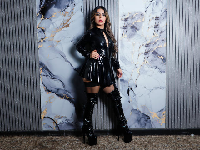 NicolBennett stands in black latex dress, high-heel boots, long wavy hair, red lipstick, showing her ass, against marble-patterned wall, with gray vertical panel background.