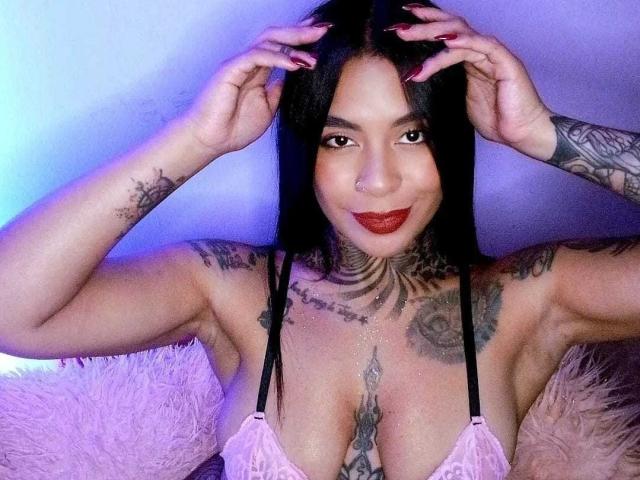 MissRuuby with black hair, tattoos, and red lipstick, smiling with hands in her hair, wearing a pink lace bra, showcasing her large tits and tattoos.