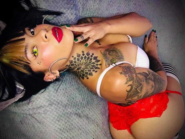 MissRuuby with tattoos, green eyes, red lipstick, white bra, red lace panties, green nail polish, lying on bed, finger on cheek, black and blonde hair, visible ass.