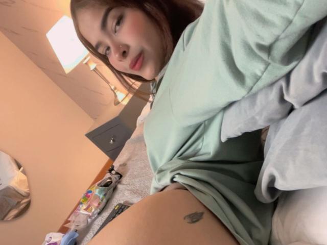 ChloeVixen con una camisa verde claro, una cama gris, un tatuaje visible en el muslo, piernas ligeramente abiertas, cabello castaño, labios rosados, dormitorio, una lámpara en el fondo. Su vulva no es visible..