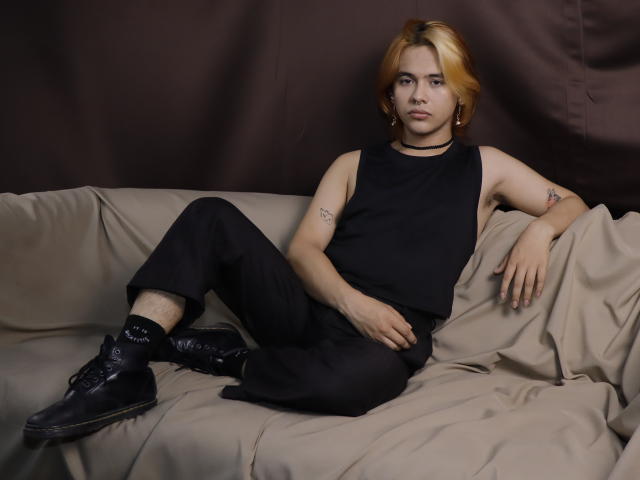 BenjiVampire with blonde hair, sleeveless black top, black pants, and black boots, lounging on a beige couch, showing tattoos on their right arm. BenjiVampire's ass is partially visible.