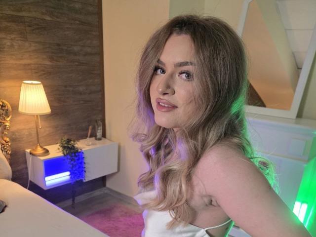 NayaQuinn has long, wavy blonde hair, smiling with subtle makeup, wearing a white strapless top, showing her shoulder and upper chest, with green and blue lights in the background.
