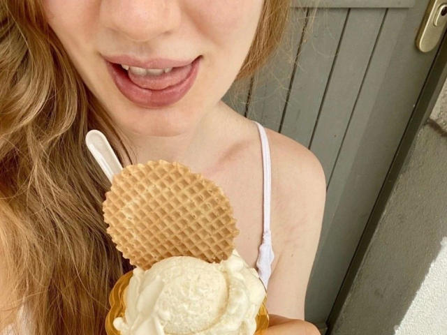 GeißeNachbarin-hot with tongue out, holding a waffle cone with ice cream and a cookie, white spoon in hand, wearing a white strap top, her tits partially visible.