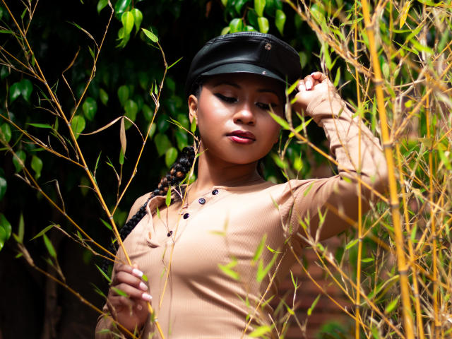EbonyxLust with dark skin, braided hair, wearing a black cap and tan button-up shirt, standing in bamboo, hand in cap, looking down, soft light, natural setting.