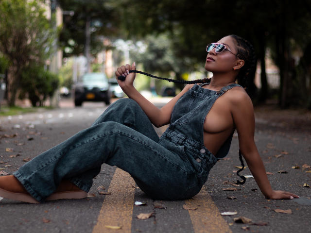 EbonyxLust sitting on a road, wearing blue denim overalls with side boob, holding a braided black string, wearing sunglasses, barefoot, with brown skin and long braids.