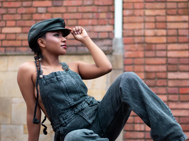 EbonyxLust, à la peau foncée, vêtue d'une salopette en jean, d'une casquette et avec des tresses, est assise contre un mur de briques, une main sur sa casquette, regardant sur le côté, montrant ses petites seins et ses fesses galbées..