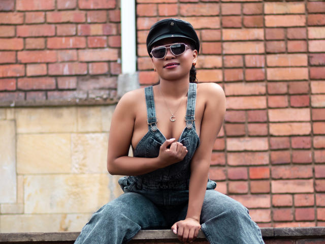 EbonyxLust, portant une casquette noire, des lunettes de soleil et une salopette en jean bleu, expose ses seins avec sa main, assise contre un mur de briques. Elle sourit et a un air mignon et sexy..