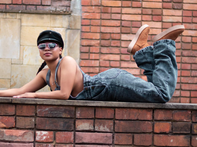 EbonyxLust ligt op een bakstenen muur, met een zwarte pet, zonnebril, een overall van denim en bruine schoenen, haar billen omhoog en haar borsten naar beneden, en ze ziet er cool en ontspannen uit..