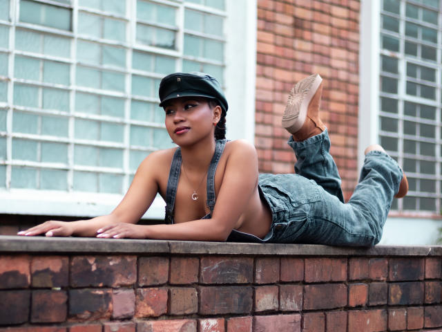 EbonyxLust, sem blusa, com seios pequenos, vestindo jeans cinzas, boné preto e botas marrons, deitada em um parapeito de tijolos, com as pernas levantadas, em frente a uma janela de blocos de vidro, olhando para o lado..
