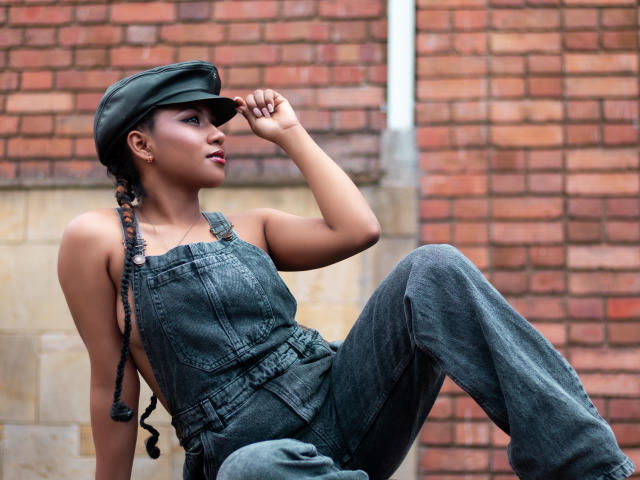 EbonyxLust, avec ses cheveux noirs tressés, portant une casquette verte, une salopette en jean et des bottes, assise contre un mur de briques, une main sur sa casquette, regardant sur le côté, mettant en valeur ses fesses..