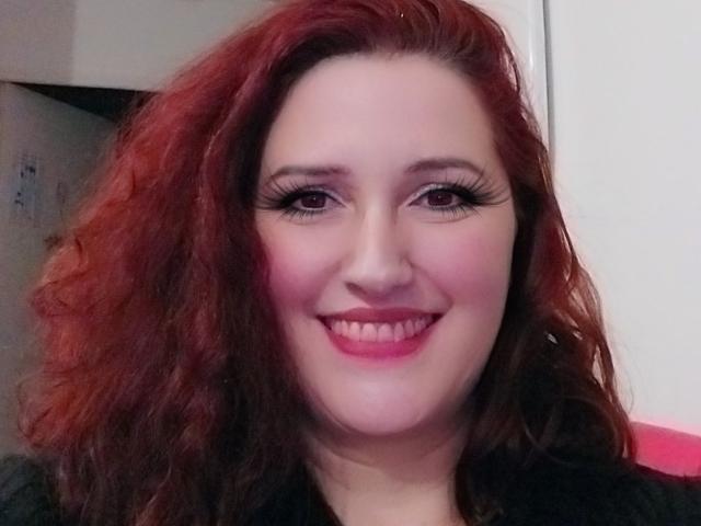 Portrait of LiliChattePoilue with curly, shoulder-length red hair. She has fair skin, pink blush on her cheeks, and thick black eyeliner with long, curved lashes. Her pink lipstick contrasts with her black top. She's smiling with visible teeth. The image is centered on her face, with her head slightly tilted to the left. The background is blurred, focusing attention on her facial features. The lighting is even, highlighting her red hair and pink blush.