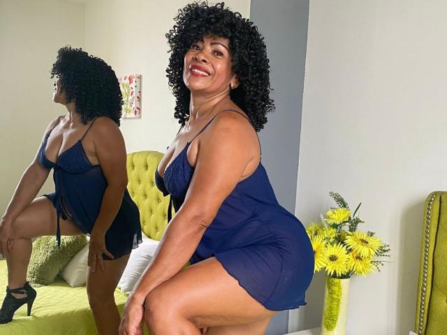 MaraRoberts, curvy black woman with curly hair, wearing a blue sheer dress, smiling, kneeling, showing her ass, reflected in a mirror, yellow flowers in a vase beside her.