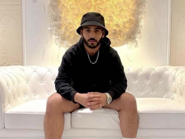 Annisse, portant un chapeau noir, un sweat à capuche noir et sans pantalon, assise sur un canapé blanc capitonné, les mains sur les genoux, portant un collier et une montre..