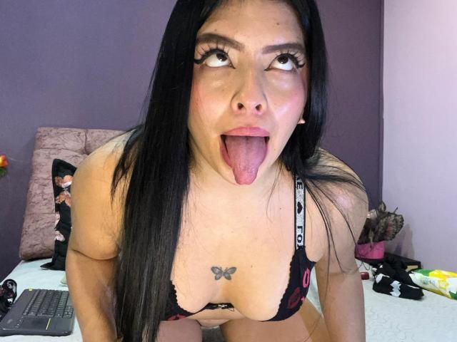 KateWelsin with long black hair, big brown eyes, and long eyelashes, tongue out, wearing a black lace bra, showing her large tits and butterfly tattoo on her chest.