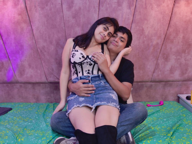 SofiNdLogan, with small tits in a floral bra, sits on a guy's lap. He's holding her ass, she's wearing a denim skirt and black socks. Purple light in background.