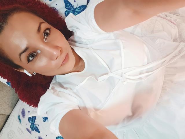 AmberDemons with red hair, white shirt, and earbuds, lying on bed, looking up, showing small tits, subtle makeup, white skin, soft light, red pillow, blue butterfly pillowcase.