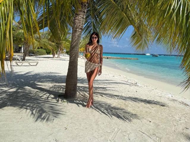 SammiMonroe stands barefoot in the sand, wearing a bikini, holding a green drink, under a palm tree with sun shadows on her tan body and dark sunglasses.