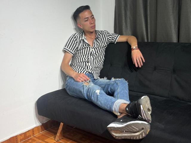 BigDickTop69 with black striped shirt, ripped blue jeans, and black sneakers, lounging on black sofa, eyes half-closed, left arm on sofa, right hand on lap.
