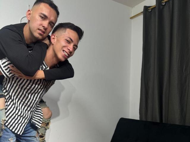 Two young men, BigDickTop69 in front and BigDickTop69 in back, standing in a room. The front BigDickTop69, with short black hair and a smile, wears a black and white striped shirt and distressed jeans. The back BigDickTop69, with short dark hair and a serious expression, wears a black long-sleeve shirt and holds the front BigDickTop69 from behind. Dark curtains on the right. A black couch on the lower right. The composition is casual and intimate.