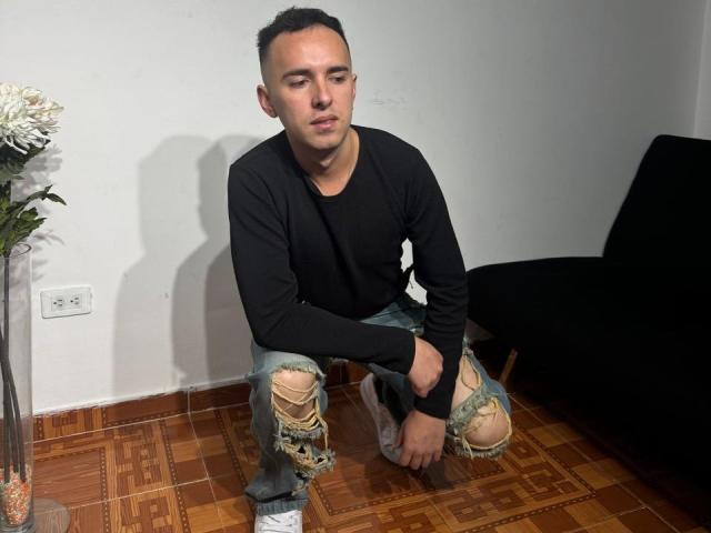 BigDickTop69 is a dude with short black hair, wearing a black long-sleeve shirt and ripped blue jeans, kneeling on wooden floor, looking down with a serious expression.