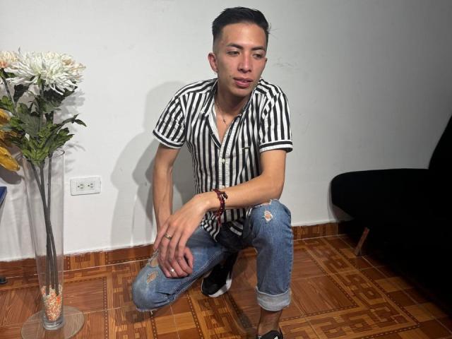 BigDickTop69, slim, wearing a striped shirt and ripped jeans, crouching on wooden floor, black chair on right, vase with white flowers on left, wrist bracelet, short dark hair.