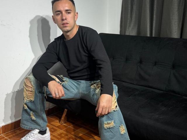 BigDickTop69 with short dark hair, black long-sleeve shirt, and ripped blue jeans, sitting on black sofa, white sneakers, wooden floor, shadow on white wall.